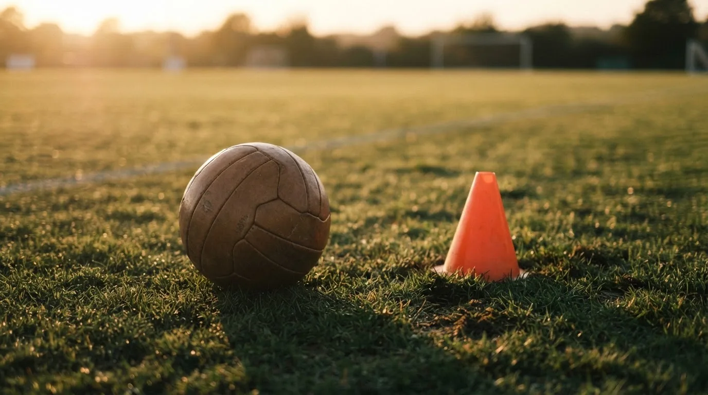 Ein Fußball liegt auf einem Trainingsplatz neben einem orangen Hütchen, Abendlicht fällt flach über den Rasen.