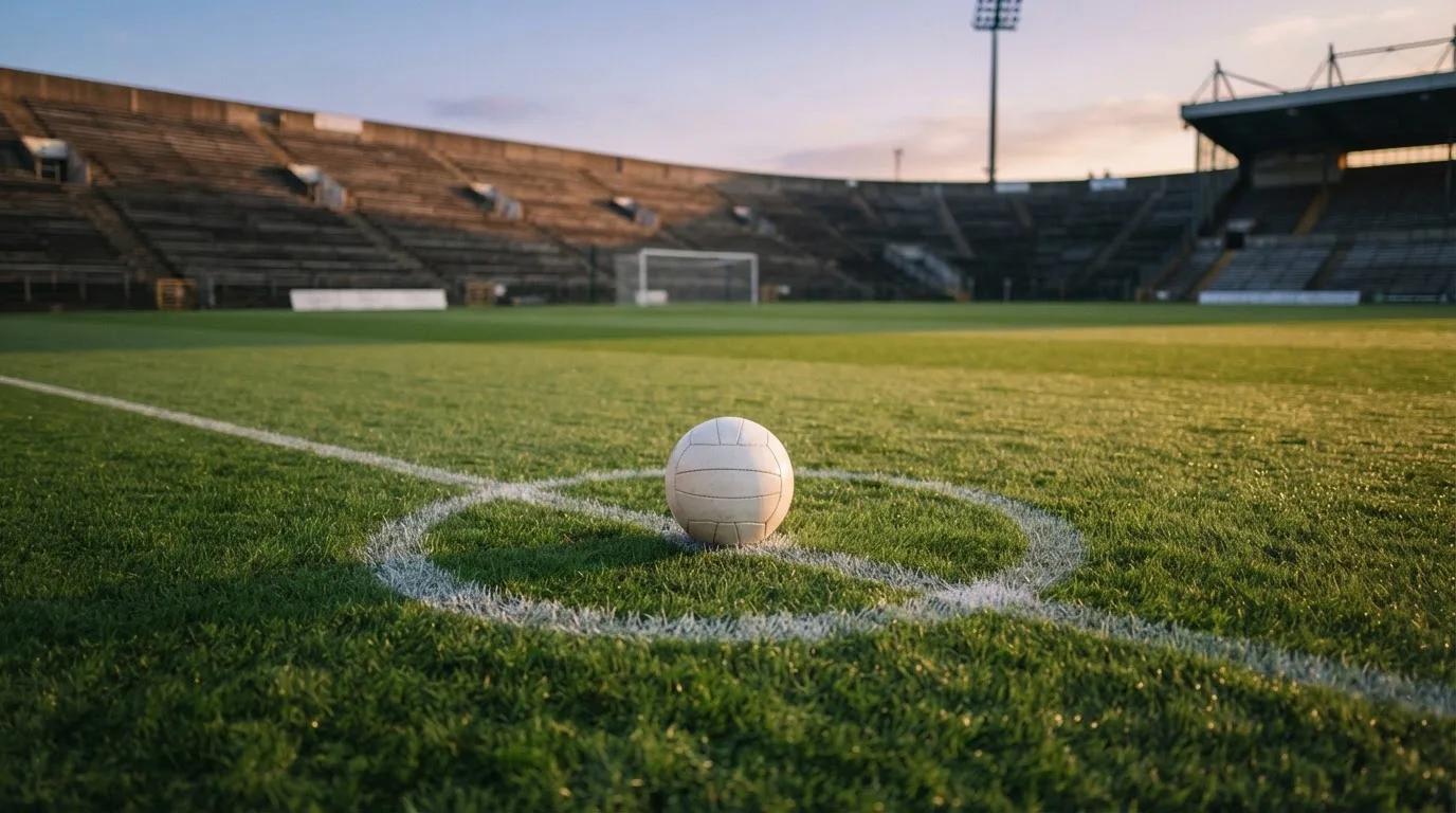Ein Fußball liegt auf dem Anstoßpunkt eines leeren Rasenplatzes, dahinter eine menschenleere Tribüne im weichen Abendlicht.