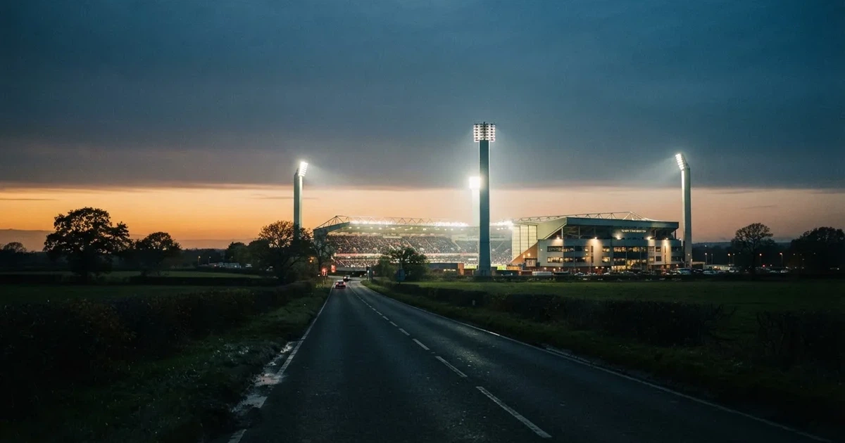Ein Stadion unter Flutlicht aus großer Entfernung gesehen, davor eine dunkle Landstraße und einzelne Bäume in der Abenddämmerung.