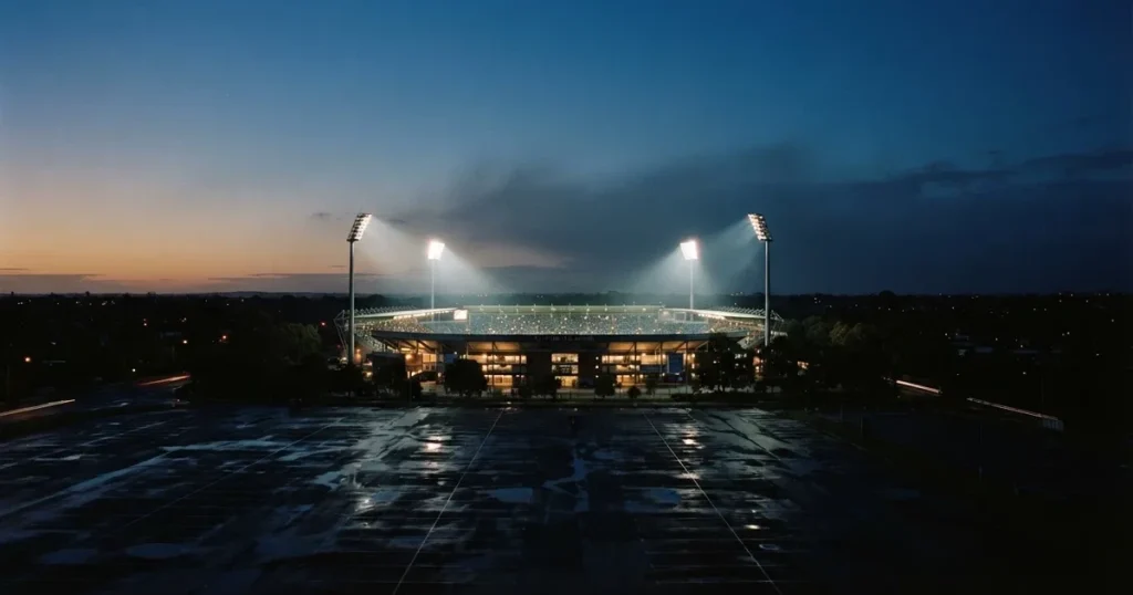 Ein Fußballstadion unter Flutlicht aus großer Entfernung gesehen, die Lichtkegel stehen im dunklen Abendhimmel über leeren Parkplätzen.