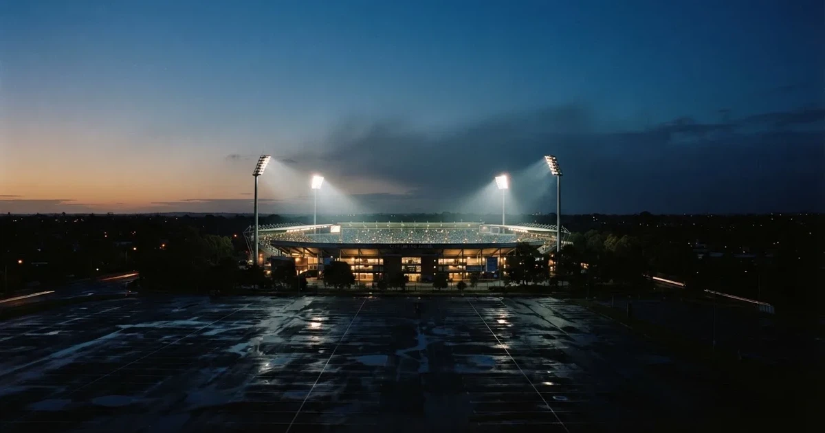 Ein Fußballstadion unter Flutlicht aus großer Entfernung gesehen, die Lichtkegel stehen im dunklen Abendhimmel über leeren Parkplätzen.