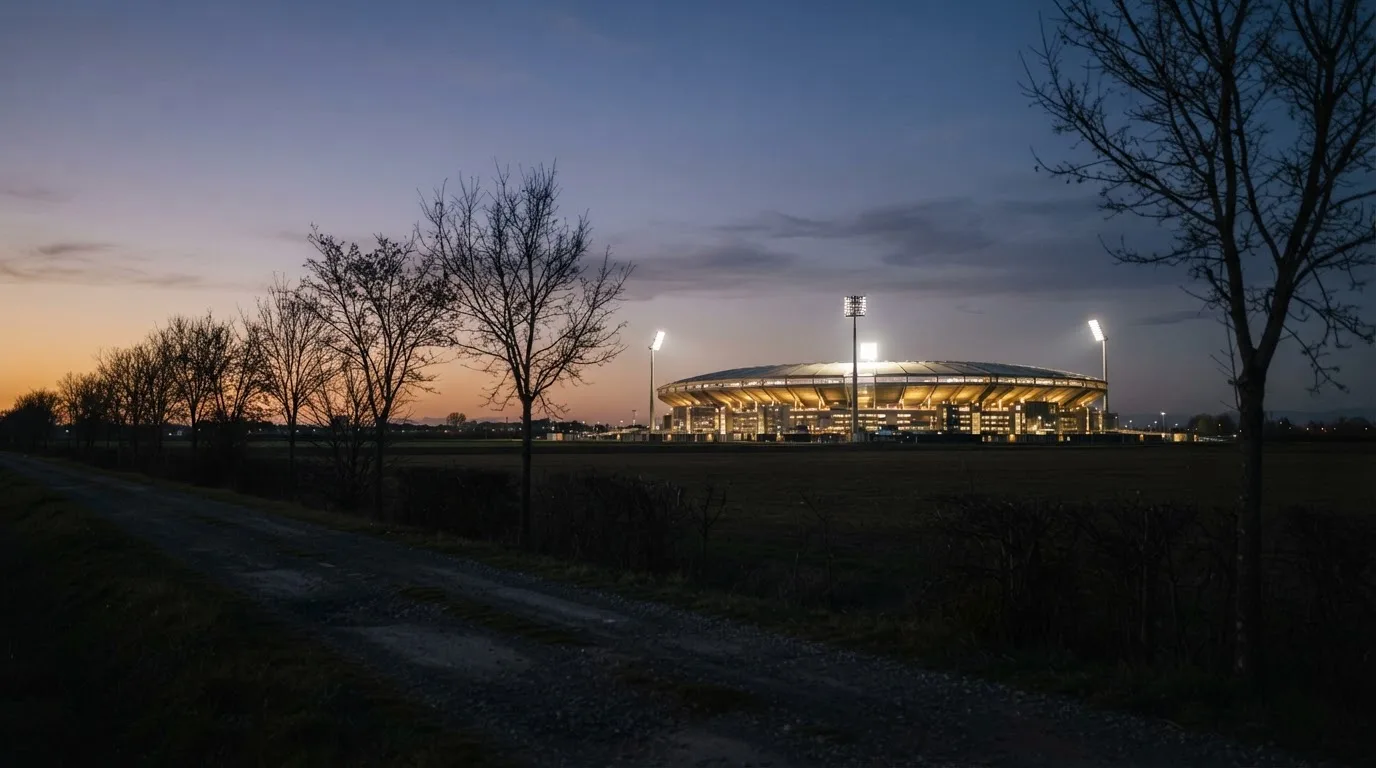 Ein großes Stadion unter Flutlicht aus weiter Entfernung gesehen, davor eine dunkle Landstraße und ein paar Bäume.