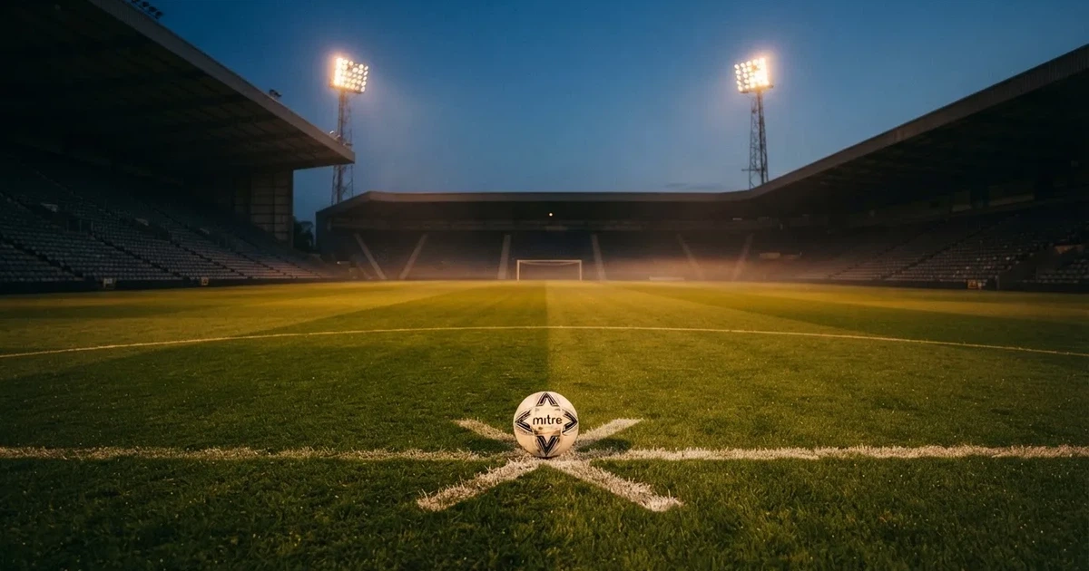 Ein Fußball liegt auf dem Anstoßpunkt eines Rasenplatzes in der Dämmerung, in der Ferne leuchten Flutlichter, die Ränge sind leer.